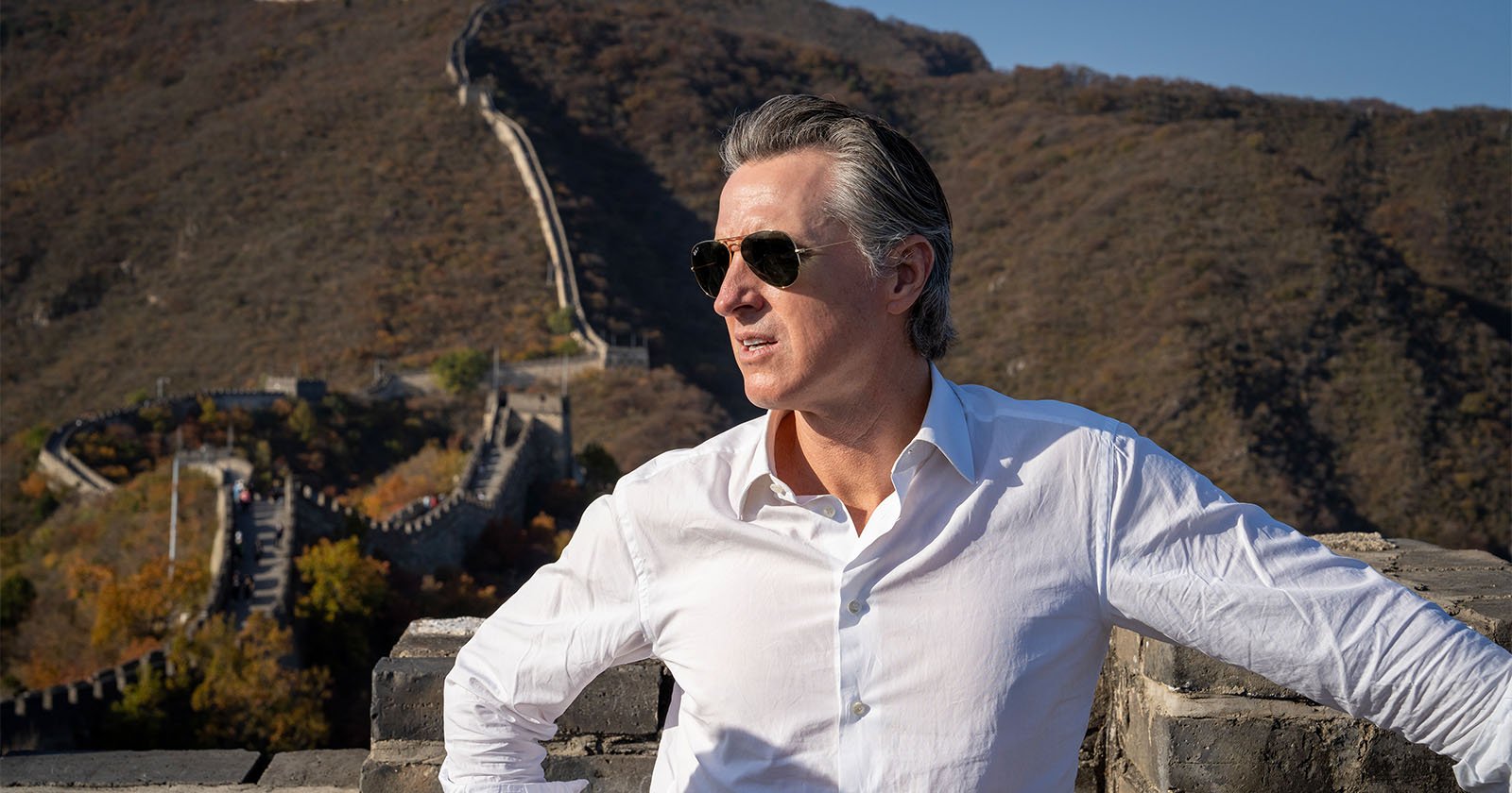 A man with gray hair and wearing sunglasses and a white shirt stands in front of a section of the Great Wall of China. The ancient structure stretches across rolling hills covered in autumn foliage under a clear blue sky.