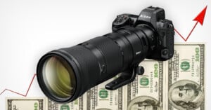 A Nikon camera with a large telephoto lens is placed on top of several hundred-dollar bills. An upward-pointing red arrow in the background suggests an increase in cost or value.