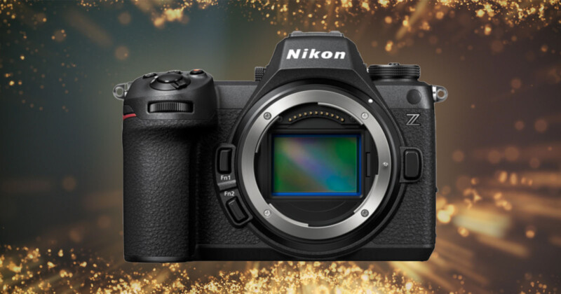 A Nikon Z camera body is photographed against a background of glowing golden lights. The camera, with its textured black exterior, prominent grip, and various control dials, has its lens mount exposed, revealing the internal sensor.