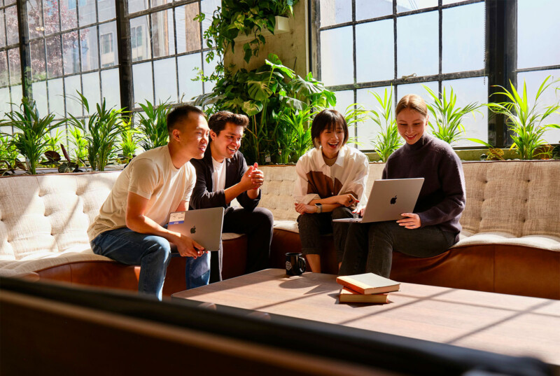 Four people are sitting on a cozy sofa near a large window, engaging in a lively discussion. Two have laptops on their laps, and all are smiling. The room is bright with natural light, and green plants are lining the window sill, adding a fresh ambiance.