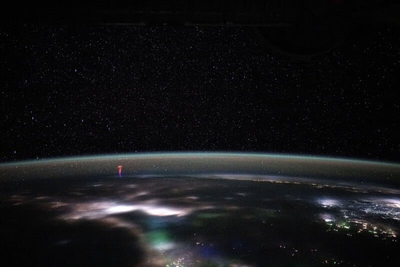 A view of the Earth from space during nighttime, showcasing a vast starry sky and a faintly illuminated horizon. Various multi-color lights and clouds are visible, with a striking red flash emerging from a cloud formation near the horizon.