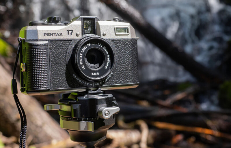 A vintage Pentax 17 film camera is mounted on a tripod with a nature background. The camera has a black textured body with silver accents and a black lens. The background includes out-of-focus elements like trees and possibly water.