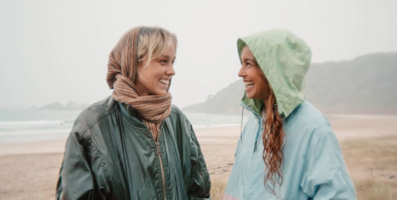 Two people wearing rain jackets and scarves are smiling and facing each other on a misty beach. The person on the left is in a dark green jacket and the person on the right is in a light green jacket. The background features a foggy shoreline and distant hills.