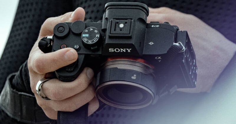 A person is holding a Sony Alpha 7 camera with both hands. The camera has a lens attached, and the person's fingers are adjusting the settings on the top dials. The background is out of focus, emphasizing the camera. The person is wearing a dark sleeve and a ring.