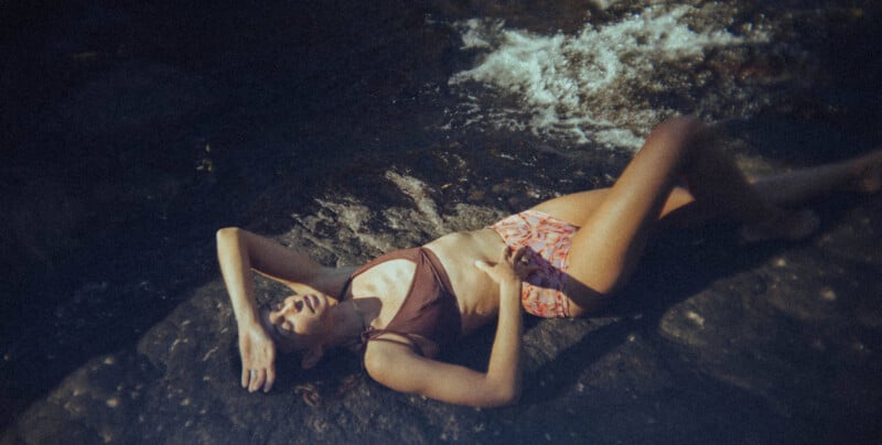 A woman wearing a bikini relaxes on a rock by a stream, with her right arm shielding her eyes and her left hand resting on her stomach. Sunlight illuminates her body while the water flows gently nearby.