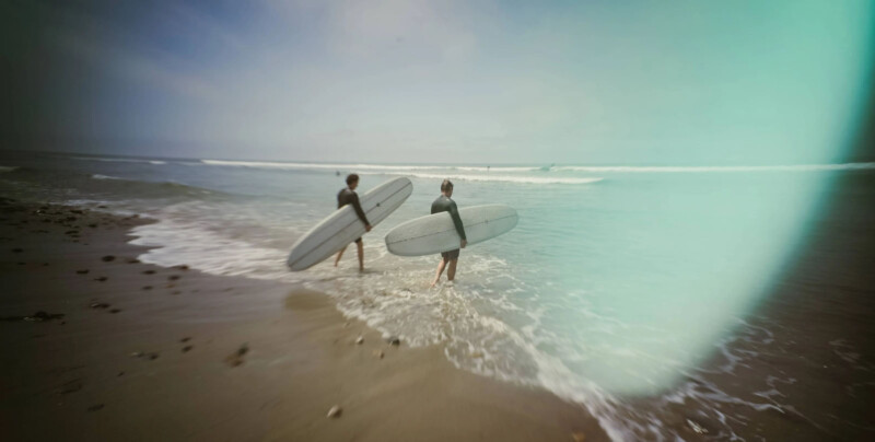 Two surfers carrying longboards wade into the ocean from a sandy beach under overcast skies. The beach has small waves approaching the shore, and the image has a greenish-blue tint. The scene captures an early morning or late afternoon surf session.