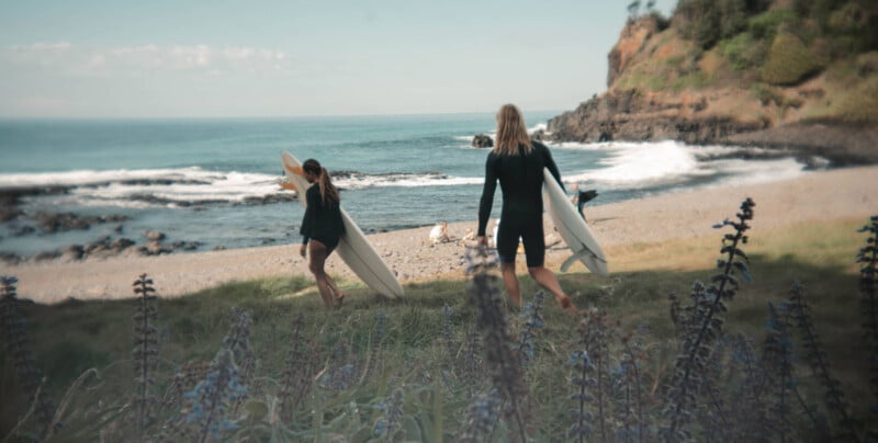 Two people carrying surfboards walk toward a rocky beach with waves crashing in the background. The shoreline is framed by a coastal cliff on the right and plants in the foreground. The sky is clear, and the scene exudes a tranquil, adventurous vibe.