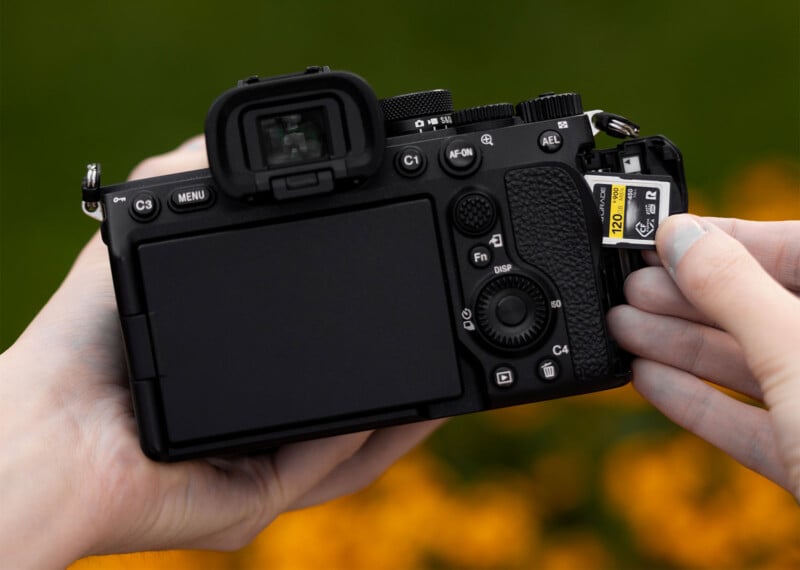 Close-up of hands inserting a 128GB memory card into the slot of a black DSLR camera. The camera's screen and various buttons are visible. The background is blurred with a mix of green and orange colors.