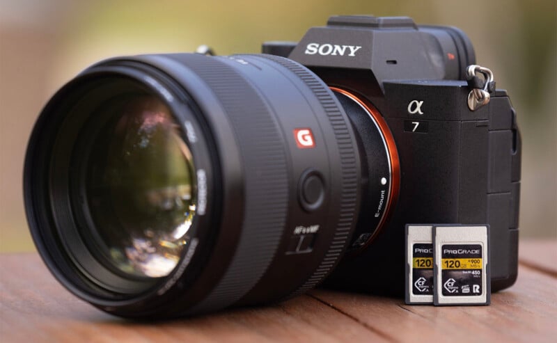 A close-up of a Sony α7 camera with a large lens attached, placed on a wooden surface. Next to the camera are two ProGrade memory cards labeled 120 GB. The background is blurred, drawing focus to the camera and memory cards.