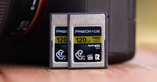 Two ProGrade 120 GB CFexpress memory cards are standing side by side on a wooden surface in front of a camera lens. The cards have a read speed of 900 MB/s and a write speed of 450 MB/s. The labels on the cards display these specifications prominently.