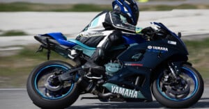 A motorcyclist in full racing gear and helmet is riding a blue Yamaha sports bike on a racetrack. The rider is leaning into a turn, demonstrating an aggressive racing posture. The background is slightly blurred, indicating high speed.