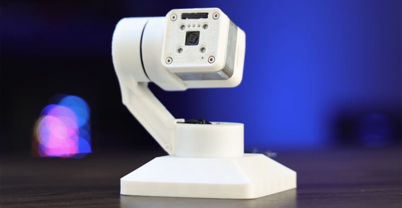 A white robotic camera mounted on a white triangular base with a swiveling arm, set against a blurred blue and black background with some colorful bokeh effects. The camera has a rectangular head with a lens in the center.