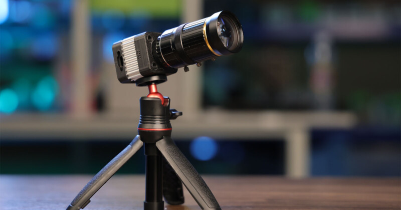 A high-quality camera with a large lens is mounted on a compact, sturdy tripod standing on a wooden surface. The background is blurred, highlighting the camera and tripod as the main focus of the image.