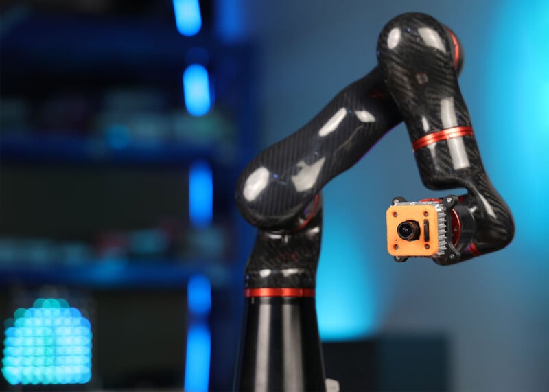 A close-up image of a robotic arm with carbon fiber segments and red connectors, holding an orange camera-like device at the end. The background is blurred, with blue lights and shelves visible.