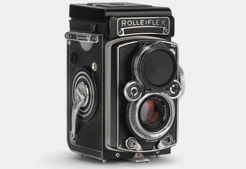 A vintage black Rolleiflex twin-lens reflex camera with silver detailing stands upright. It features a viewing lens above the taking lens, focus knobs on the side, and the recognizable "ROLLEIFLEX" brand name at the top. The camera has a classic, retro design.