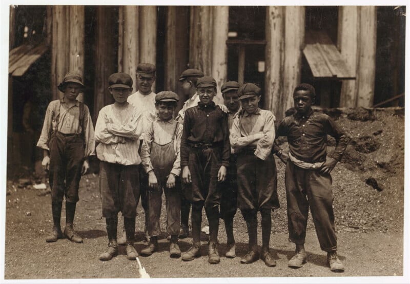 A black-and-white photo shows a group of eight young boys standing in front of large wooden beams. They are wearing work clothes, such as caps, shirts, suspenders, and boots. The boys have serious expressions and appear to be posed for the picture.