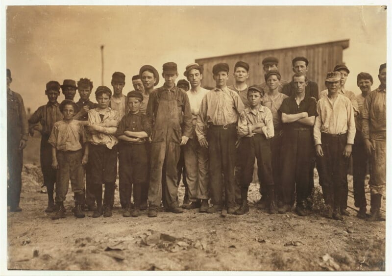 A sepia-toned photograph shows a group of boys and young men posing outdoors. They are dressed in work clothes, including overalls, hats, and long-sleeved shirts. A wooden building stands in the background. The mood appears solemn and serious.