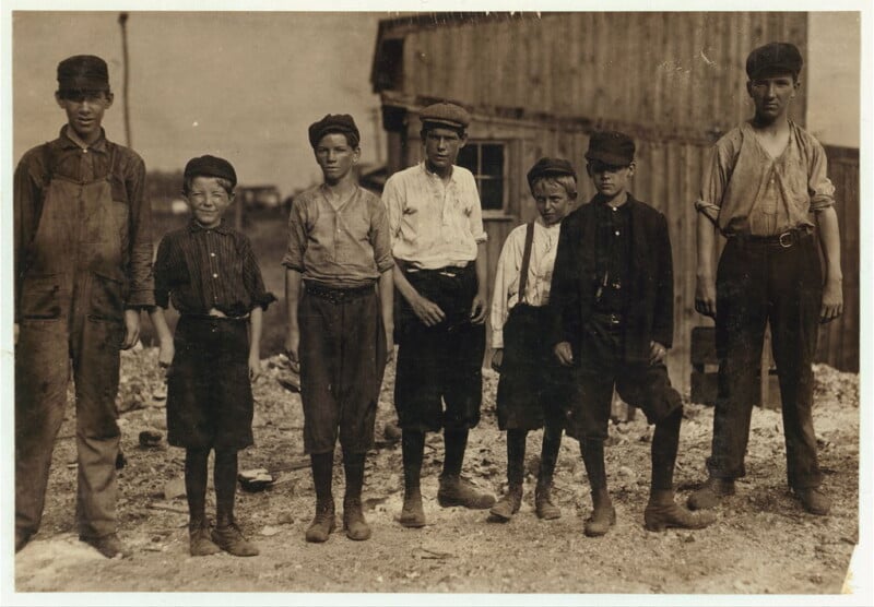 Seven young boys in work clothes, including shirts with rolled sleeves, caps, suspenders, and high boots, stand in a row with a wooden building in the background. The scene appears to be from an early 20th-century industrial or rural setting.