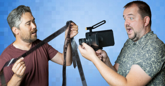 Two men are interacting with film equipment in front of a blue background. The man on the left holds a strip of film, examining it closely, while the man on the right, with a surprised expression, holds a large film camera aimed at the film strip.