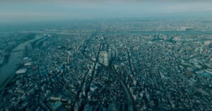 Aerial view of a sprawling urban cityscape with densely packed buildings, roads crisscrossing the landscape, and rivers cutting through the city. The horizon is hazy, indicating the vast expanse of the metropolitan area.