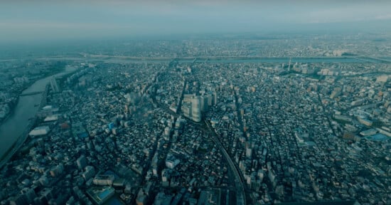 Aerial view of a sprawling urban cityscape with densely packed buildings, roads crisscrossing the landscape, and rivers cutting through the city. The horizon is hazy, indicating the vast expanse of the metropolitan area.