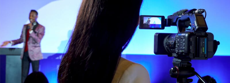 A video camera on a tripod records a person speaking on stage. The camera screen shows the recorded person, who is standing at a podium against a blue-lit background. The back of the camera operator's head is visible in the foreground.