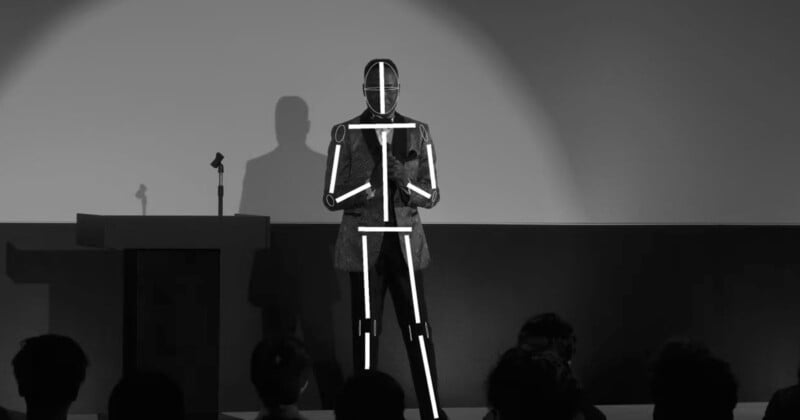 A person on stage delivers a presentation. They wear a suit with white light strips and geometric patterns, creating a futuristic appearance. There is a podium with a microphone to the left, and an audience is visible in the foreground. The image is in black and white.