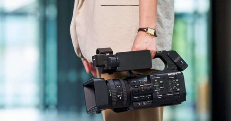 A person wearing a light-colored outfit is holding a professional Sony video camera by their side. Background is blurred with soft lighting, focusing attention on the camera.