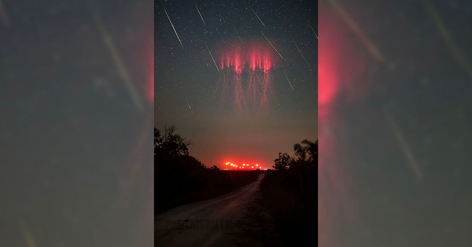 A dark, rural road stretches ahead under a night sky filled with stars and streaks of meteors. The focal point is a captivating display of red sprites lighting up the sky near the horizon, casting a surreal red glow against the landscape below.