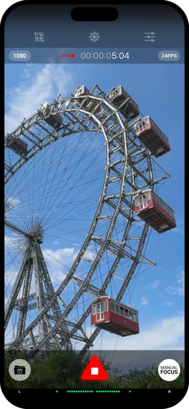 Image of a smartphone screen displaying a video recording interface while capturing footage of a Ferris wheel against a partly cloudy sky. The Ferris wheel has red gondolas, and the screen shows recording indicators and a manual focus button.