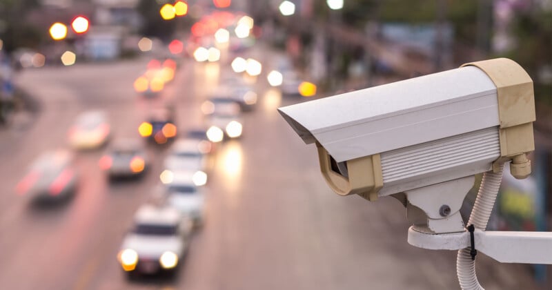 Close-up of a security camera mounted on a pole, facing a busy street. The street, out of focus, shows the blurred motion of cars and the glow of brake lights, capturing a bustling urban scene during dusk or dawn.