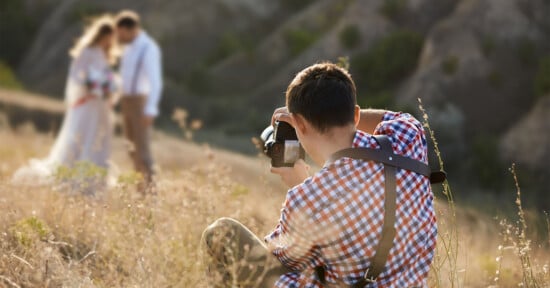 A photographer in a plaid shirt crouches in a grassy field, focusing his camera on a newly married couple in wedding attire who stand together in the blurred background against a natural, rocky landscape.