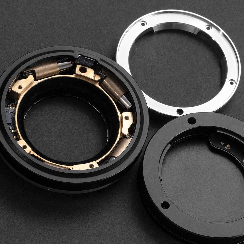 Close-up of a disassembled camera lens mount adapter. The image shows three main components: a circular black and gold component with visible electronic contacts, a silver metal ring, and a black plastic cap. All are placed on a dark, flat surface.