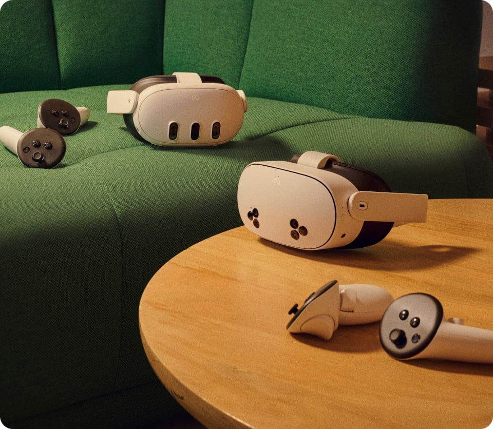 Two virtual reality headsets and four controllers are displayed. One set is placed on a green couch, while the other set is on a round wooden table in front of the couch, suggesting a relaxed, indoor setting for VR gaming or experiences.