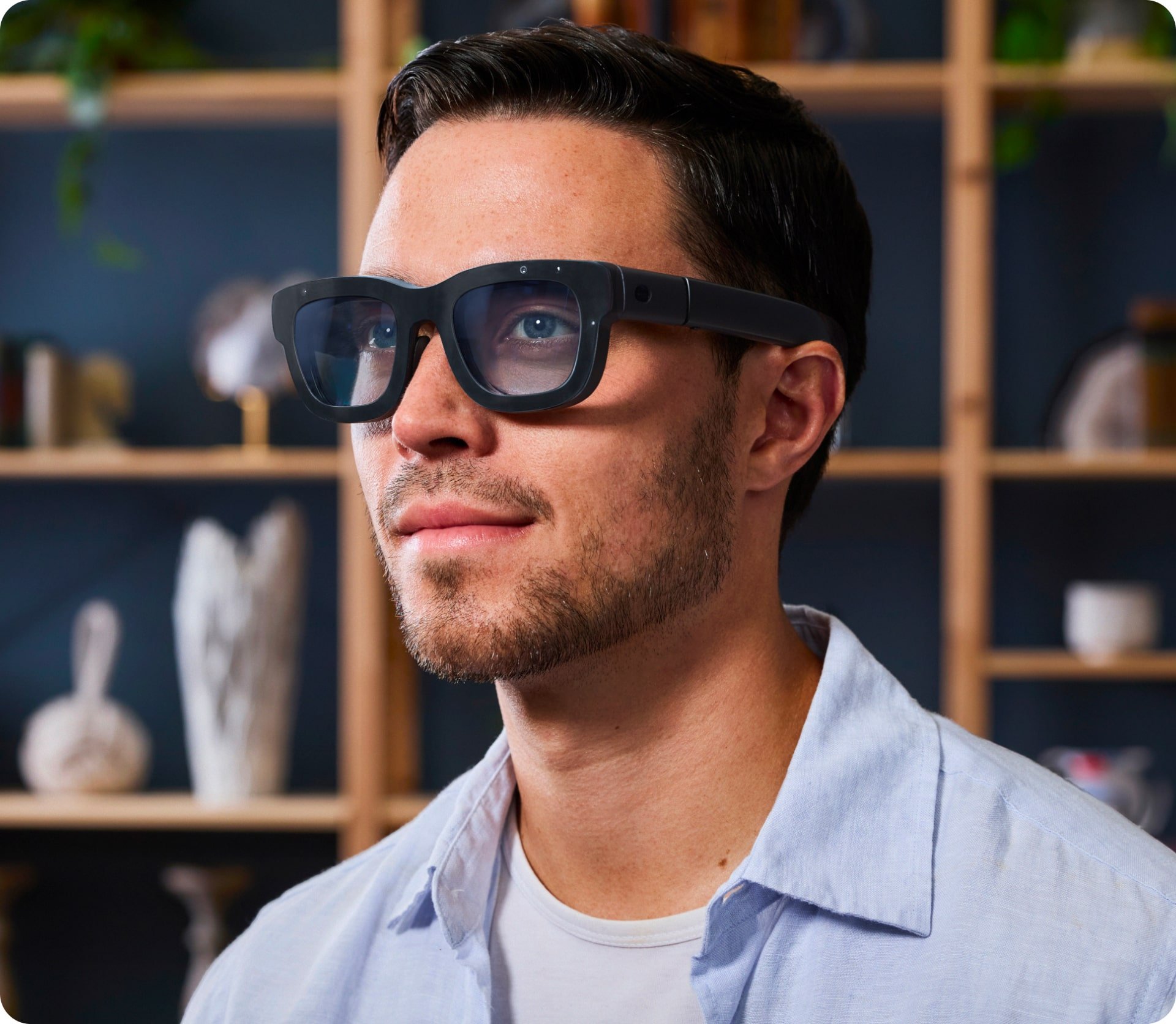 A man with short dark hair and a beard is wearing a light blue shirt and augmented reality glasses while looking slightly upward. The background is a blurred shelf with various decorative items.