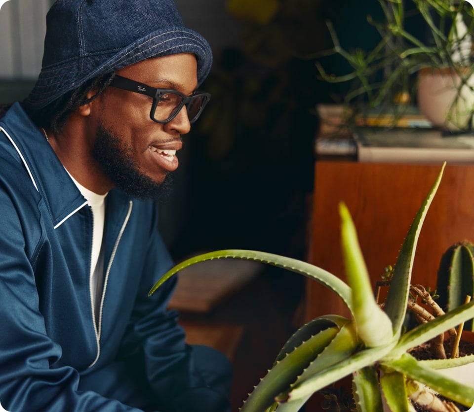 A man wearing glasses, a blue jacket, and a matching hat smiles while looking at a green aloe vera plant indoors.