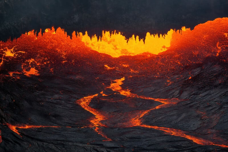 A vivid, close-up view of flowing lava, glowing bright orange as it emerges from a volcanic fissure. The molten rock contrasts against the dark, ashy surroundings, creating a dramatic scene of intense heat and geological activity.