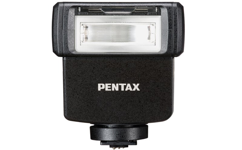 A Pentax camera flash unit with a black exterior, featuring a white flash cover and labeled "Pentax" in white text on the front. The flash has a mounting bracket at the bottom for attaching to a camera.