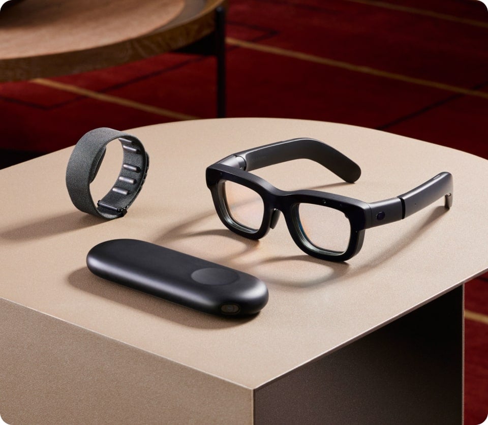 A pair of augmented reality glasses, a sleek wristband, and a compact black electronic device are displayed on a beige tabletop. The background features a brown and red patterned surface.