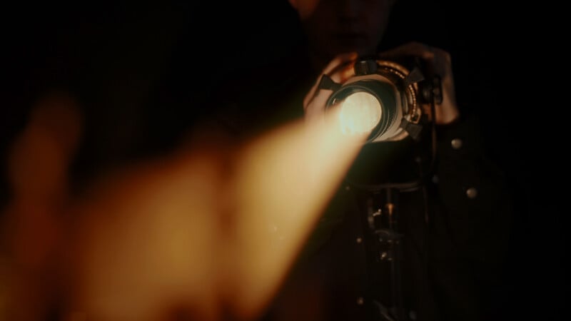 A person holding and operating a camera with a bright spotlight, casting a warm beam of light forward in a dark setting. The spotlight creates a dramatic and moody atmosphere, highlighting the intensity of the scene.