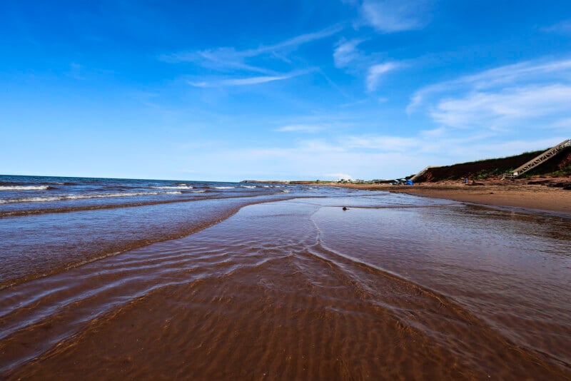 A serene beach scene under a clear blue sky, with gentle waves lapping against the sandy shore. A distant green hill, several scattered beachgoers, and a staircase leading down to the beach are visible in the background.