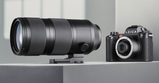 A high-resolution digital camera with and a telephoto lens on display. The camera has a sleek black design and the lens is large with prominent ridges, both placed on a minimalist white surface. The background features a soft, blurred out window light.