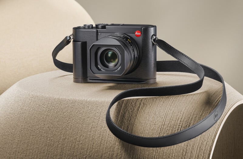 A black Leica Q2 camera with a leather strap is placed on a curved beige surface. The camera's lens is prominently displayed, and the Leica logo is visible on both the camera body and the strap. The background is soft and neutral-toned.