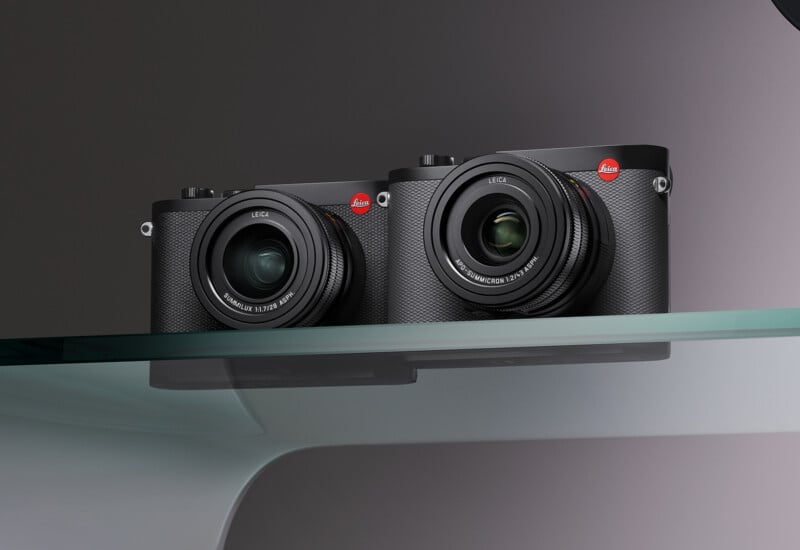 Two black Leica cameras with lenses attached are placed side by side on a glass shelf. Both cameras prominently feature the red Leica logo in the top right corner. The background is a neutral gradient.