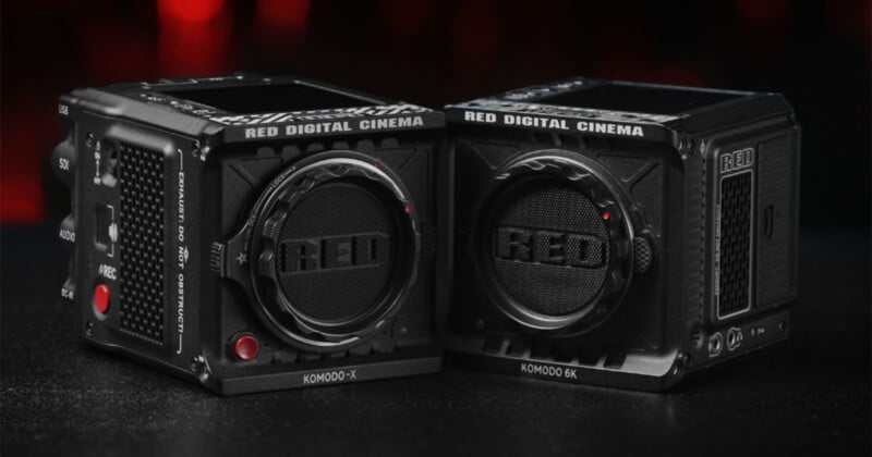 Two black RED Digital Cinema Komodo-X and Komodo 6K cameras sit side by side on a dark surface with a red and black blurred background. The lenses prominently display the "RED" logo.