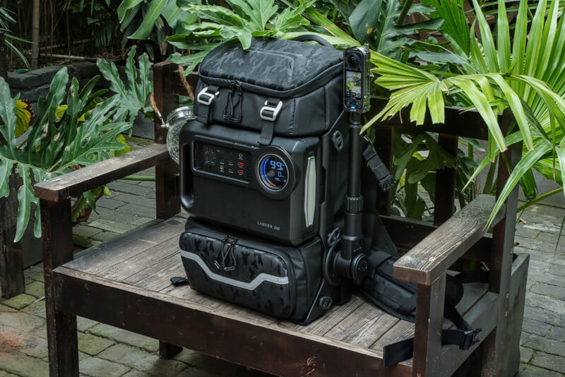 A multifunctional black backpack with electronic components is situated on a wooden bench in a lush garden. The backpack features a digital display, various buttons, and pockets, with a smartphone and tripod attached to the side.