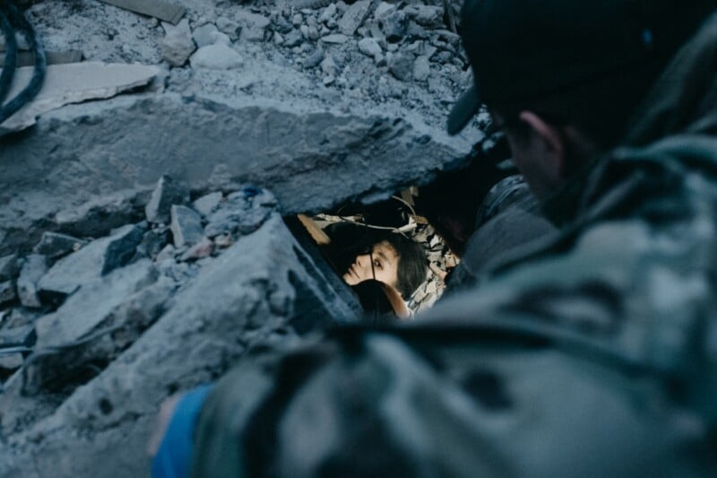 A person is seen trapped under rubble, with only their face visible through a small opening. Another individual in military or rescue clothing peers down at them, surrounded by debris and broken concrete. The scene is lit by a focused light, highlighting the person’s face.