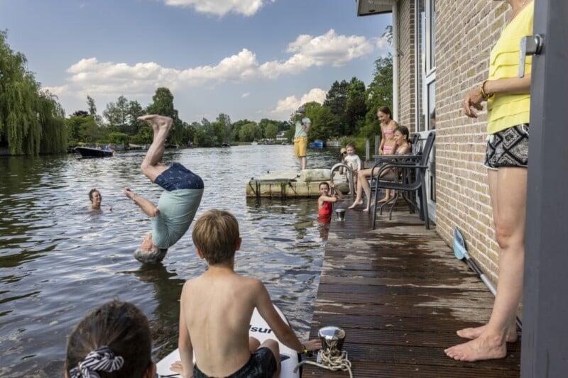 A group of people gather by a dock next to a brick house. A man is mid-flip into the water while children watch from the dock and a small boat. Some people are seated on chairs, while a woman in a yellow shirt stands nearby. Trees and boats are visible in the background.