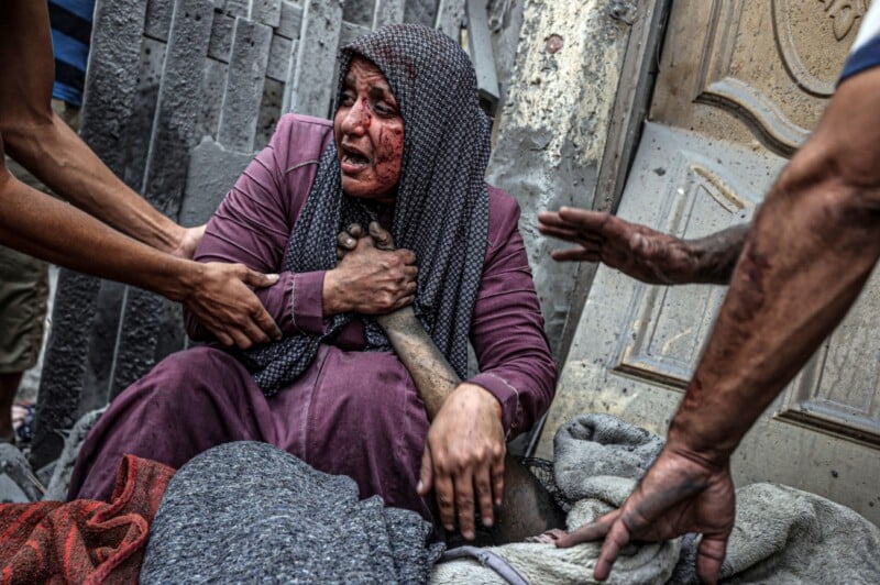 A distressed woman with a bloodied face sits on the ground, grasping a lifeless arm. She wears a purple garment and a patterned headscarf. Three individuals around her extend their hands in what appears to be an attempt to offer help. The setting appears to be chaotic and damaged.