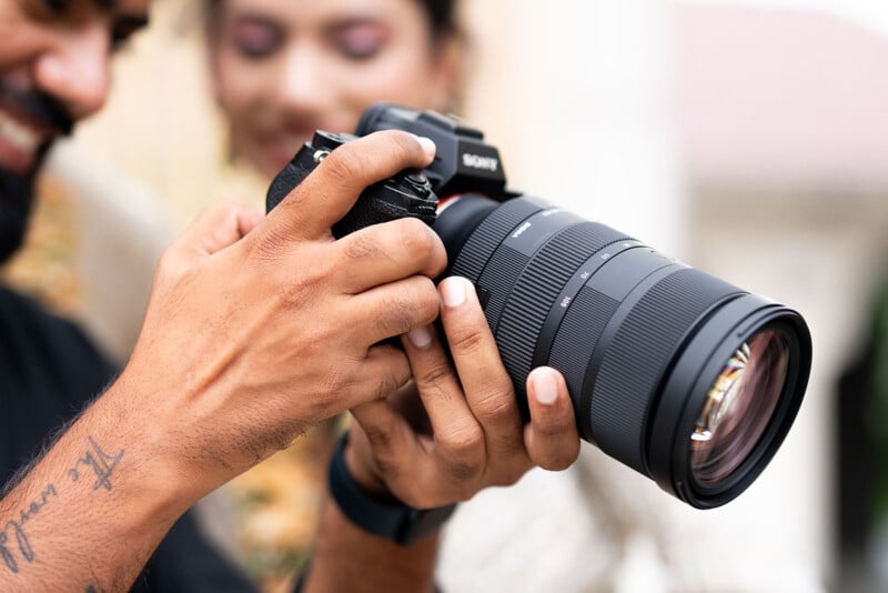 Close-up of a person holding a professional camera and adjusting its settings, with another person smiling in the background. The camera features a large telephoto lens, and the scene conveys a joyful moment of photography.